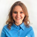 Mulher jovem com cabelos castanhos ondulados e camisa azul, sorrindo em frente a um fundo branco.
