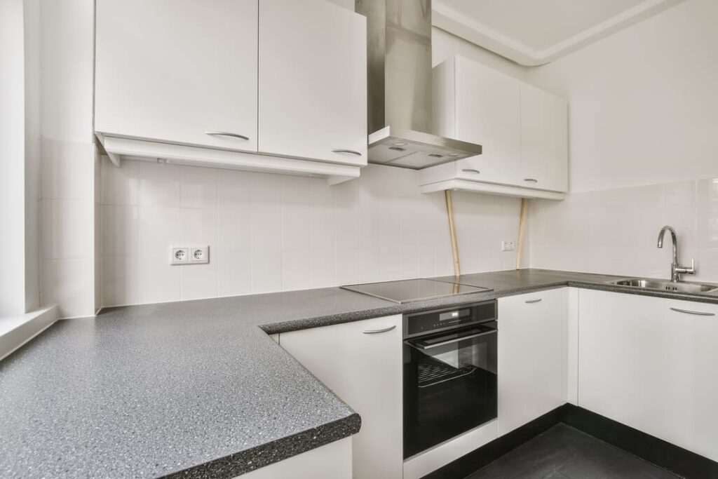 Cozinha moderna em formato L com bancada de granito cinza escuro, arm&aacute;rios brancos e coifa de inox.