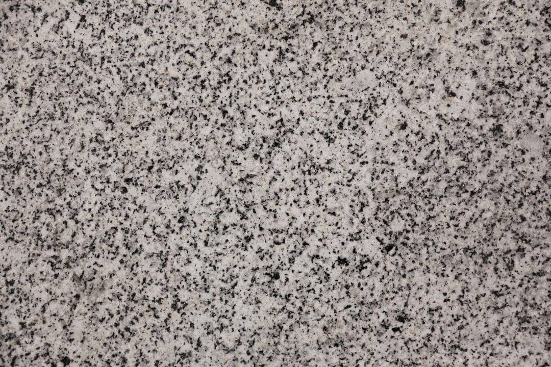 A imagem mostra uma textura de granito em close-up, destacando os detalhes das manchas pretas e cinzas sobre um fundo acinzentado mais claro. O granito apresenta um padrão natural, irregular e granulado, com pequenos pontos de cor distribuídos de forma aleatória, característicos desse tipo de pedra. A aparência é rugosa e sólida, transmitindo a sensação de durabilidade e resistência, típica de superfícies de granito.