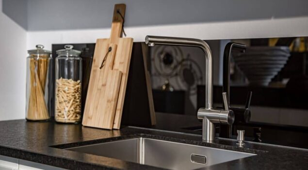 Imagem de uma pia de cozinha moderna com bancada escura e cuba de aço inox. Em destaque, uma torneira de design minimalista em acabamento metálico. Ao lado da pia, há potes de vidro com espaguete e macarrão, além de duas tábuas de madeira encostadas na parede, criando um ambiente organizado e sofisticado.