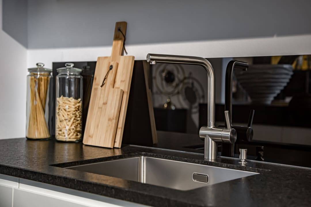 Imagem de uma pia de cozinha moderna com bancada escura e cuba de aço inox. Em destaque, uma torneira de design minimalista em acabamento metálico. Ao lado da pia, há potes de vidro com espaguete e macarrão, além de duas tábuas de madeira encostadas na parede, criando um ambiente organizado e sofisticado.