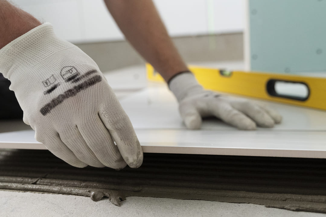 Pessoa com luvas brancas está instalando uma placa cerâmica ou pedra sobre uma superfície com argamassa. Ao fundo, há um nível amarelo, indicando o cuidado com o alinhamento.