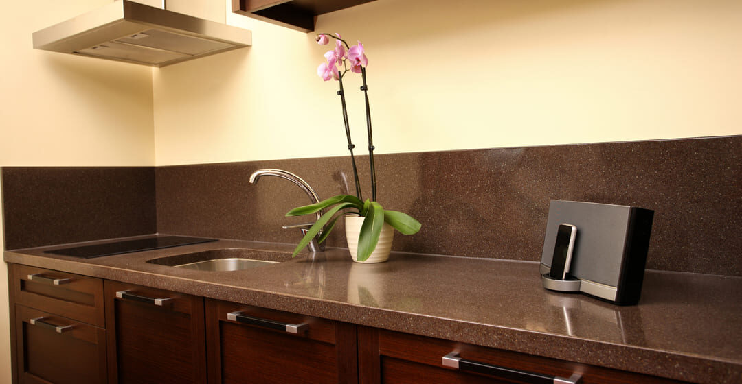 Cozinha moderna com bancada de granito marrom e armários de madeira escura. A pia embutida tem uma torneira metálica curva. Sobre a bancada, há um vaso branco com uma orquídea de flores roxas e um telefone sem fio em sua base de carregamento. No fundo, um exaustor de aço inoxidável e armários suspensos completam o ambiente.