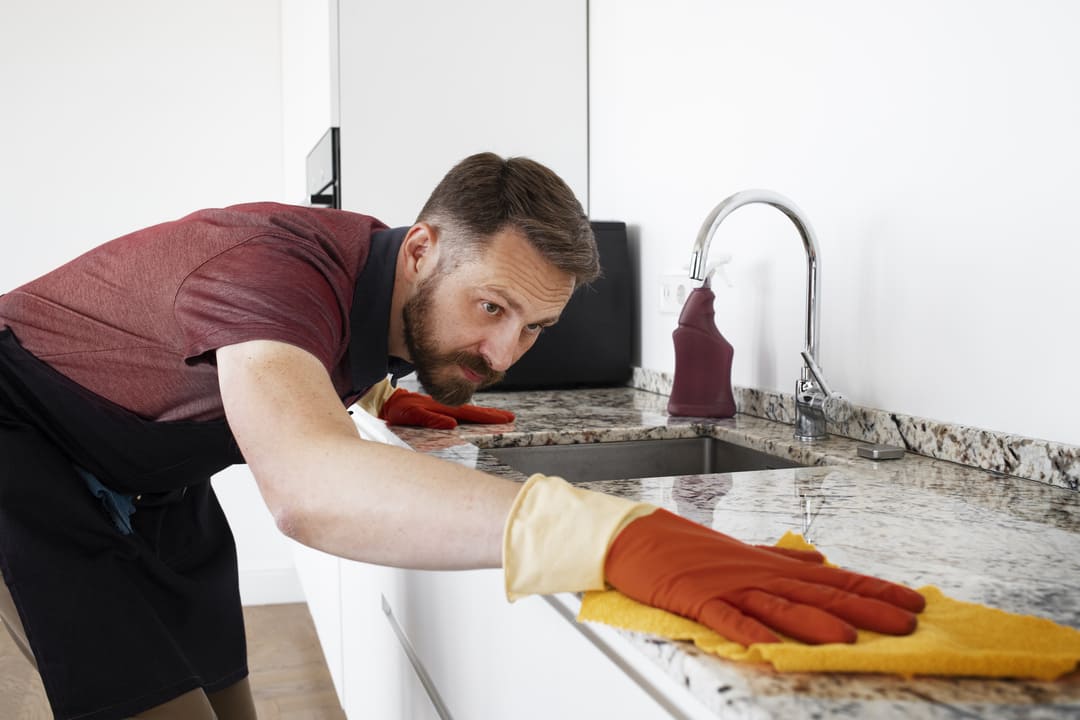 Homem usando luvas de limpeza laranja e um avental preto, limpando atentamente uma bancada de granito em uma cozinha moderna.