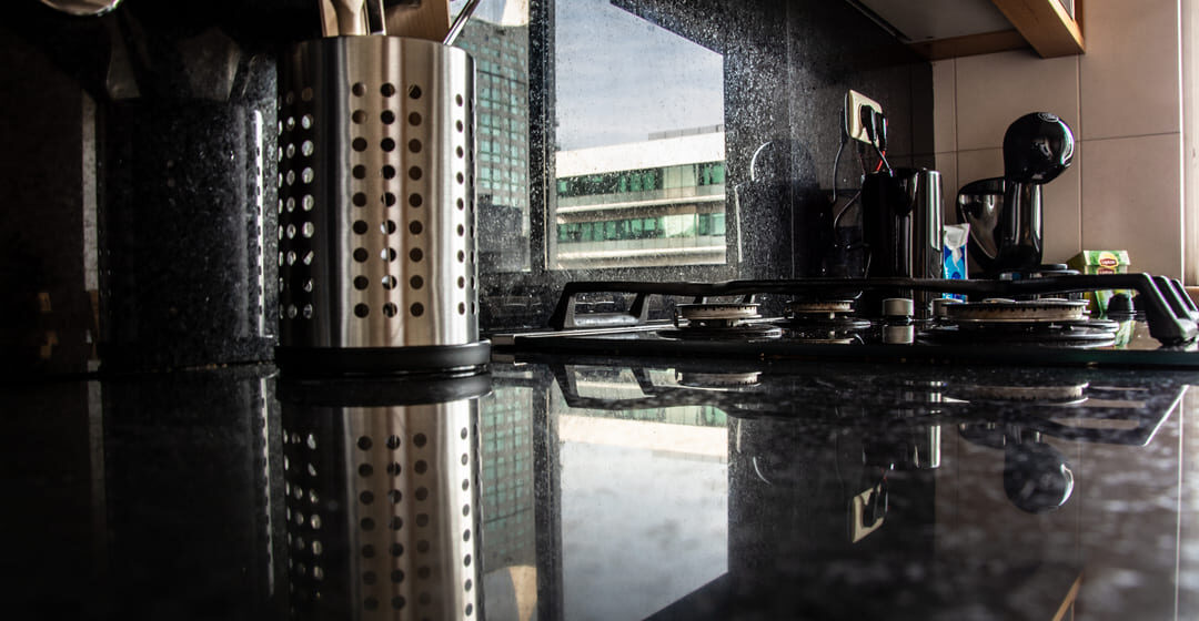 Bancada de cozinha com granito preto polido, utensílios inox e fogão cooktop refletidos na superfície.