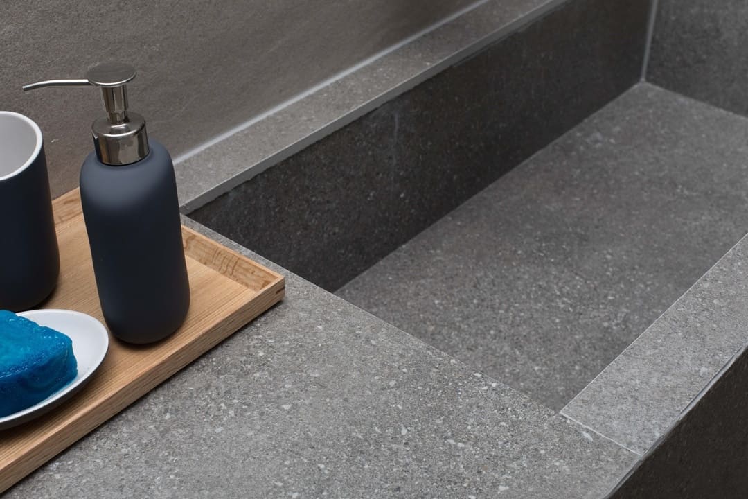 Bancada de pedra cinza com acabamento fosco, contendo uma saboneteira com sabão azul e um dispenser de sabão líquido sobre uma bandeja de madeira.