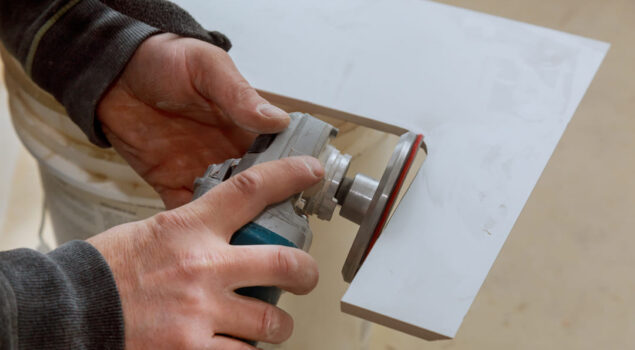 Pessoa utilizando uma politriz elétrica com disco abrasivo para lixar a borda de uma peça de mármore branco, representando o uso de abrasivos na marmoraria.