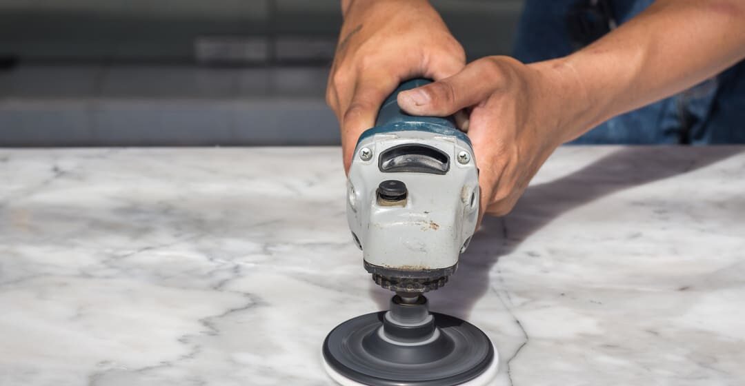 Pessoa utilizando uma politriz elétrica para polir uma superfície de mármore branco com veios cinza.