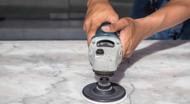 Pessoa utilizando uma politriz elétrica para polir uma superfície de mármore branco com veios cinza.