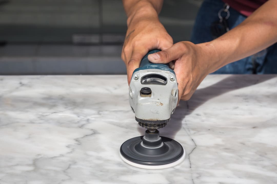 Pessoa utilizando uma politriz elétrica para polir uma superfície de mármore branco com veios cinza.