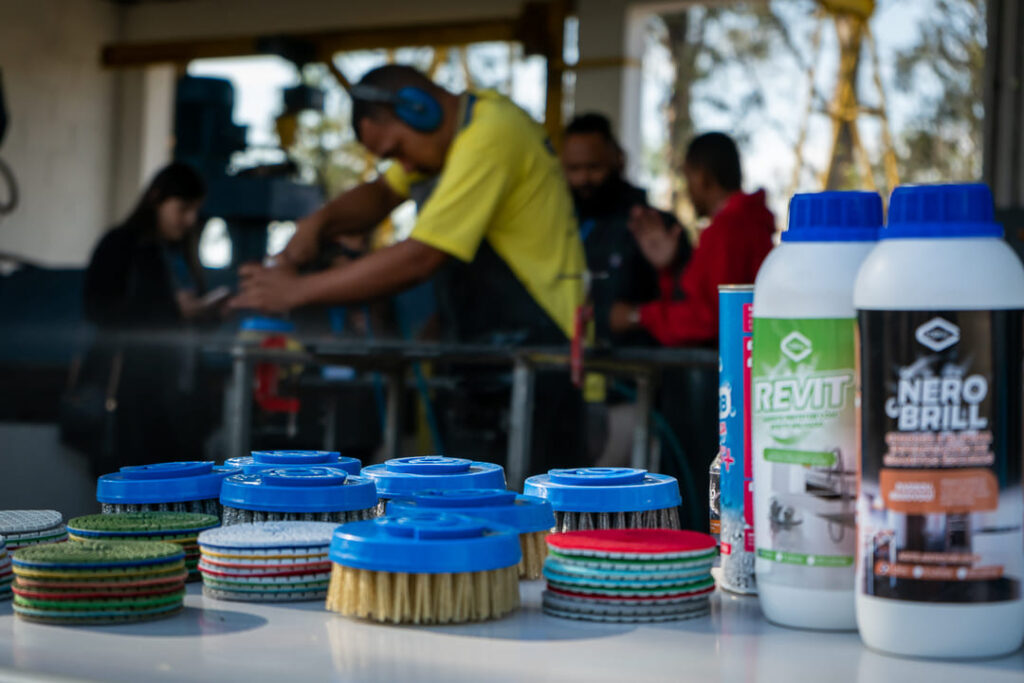 Produtos e acessórios da linha Cobral para marmoraria em primeiro plano, com operadores trabalhando ao fundo em uma bancada.