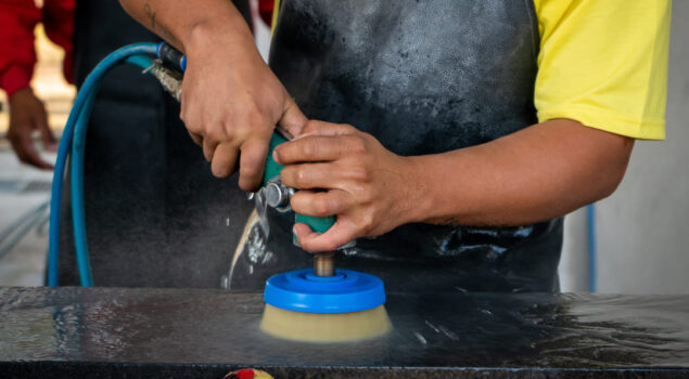 Trabalhador aplicando acabamento fosco em pedra com politriz industrial e uso de água.