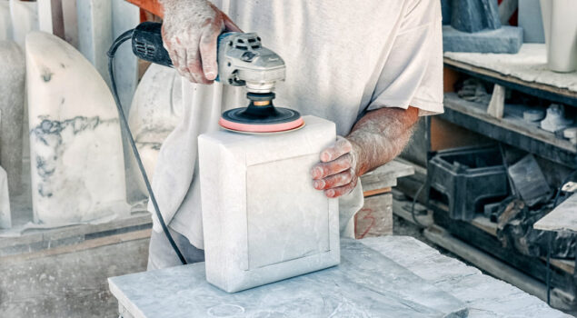 Profissional utilizando uma lixadeira elétrica para polir uma peça de mármore ou pedra, evidenciando o processo de desgaste abrasivo.
