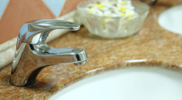 Torneira de metal cromado sobre bancada de granito marrom-claro, com uma pia branca e um recipiente de vidro contendo flores ao fundo.