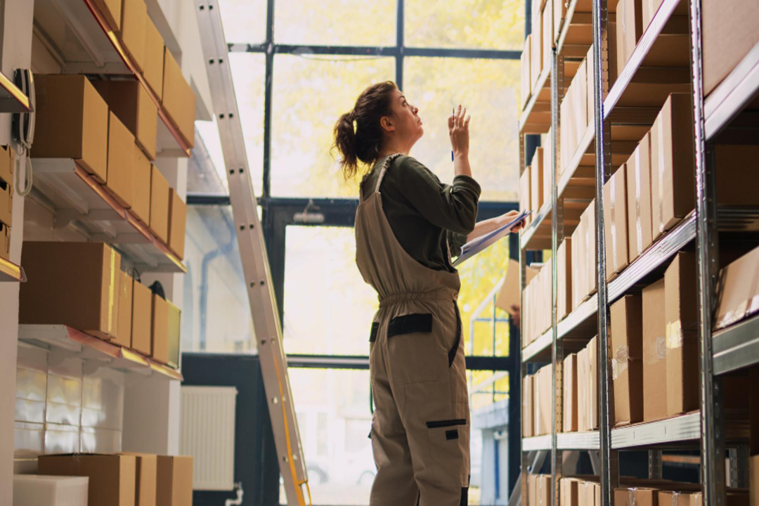 Profissional confere caixas e itens em prateleiras de um estoque organizado, utilizando uma prancheta para controle de materiais.